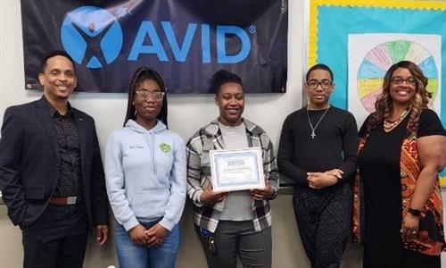 A teacher flanked by a student and staff smiles while holding her certificate.